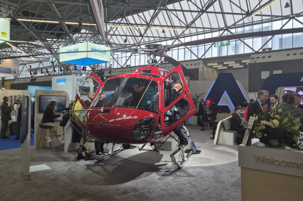 Helicóptero rojo de Air Zermatt expuesto en un pabellón ferial junto a visitantes.
