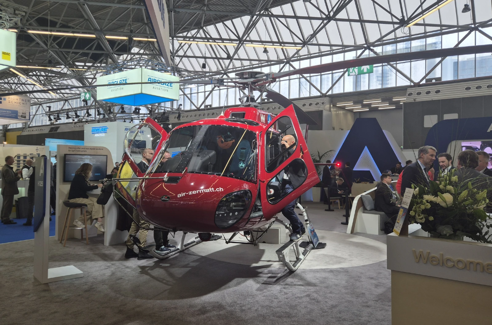 Helicóptero rojo de Air Zermatt expuesto en un pabellón ferial junto a visitantes.